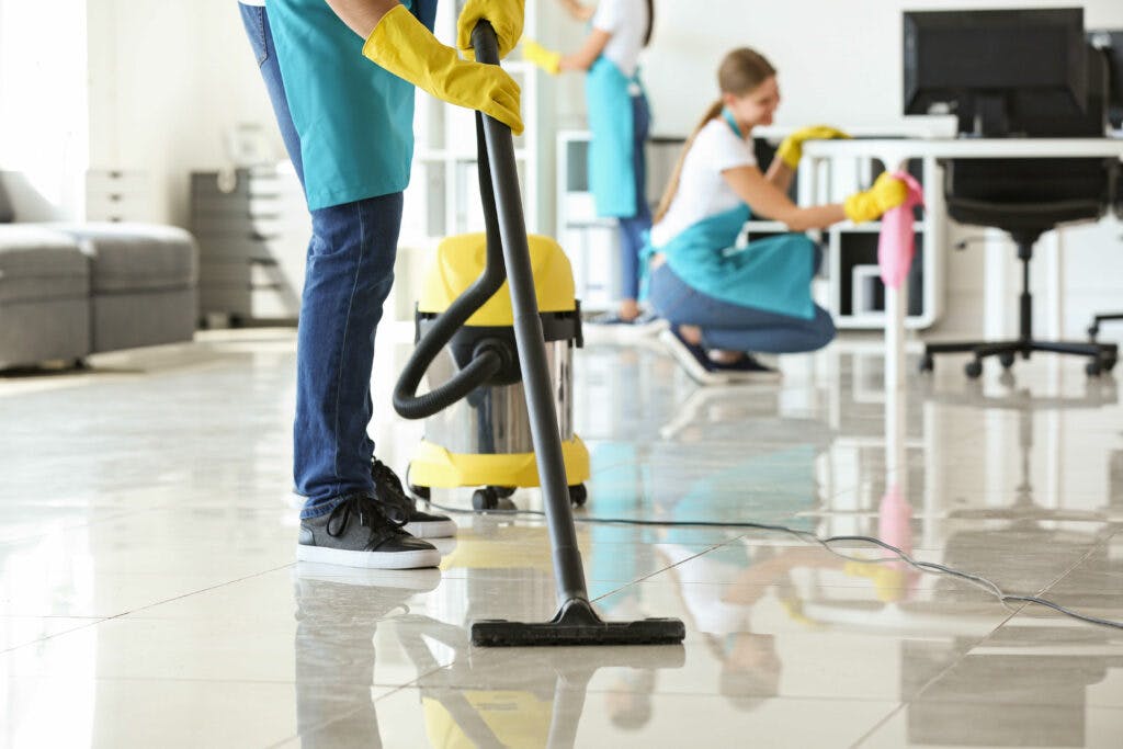 Cleaner vacuuming floor while another cleaner sanitizes a desk in the background